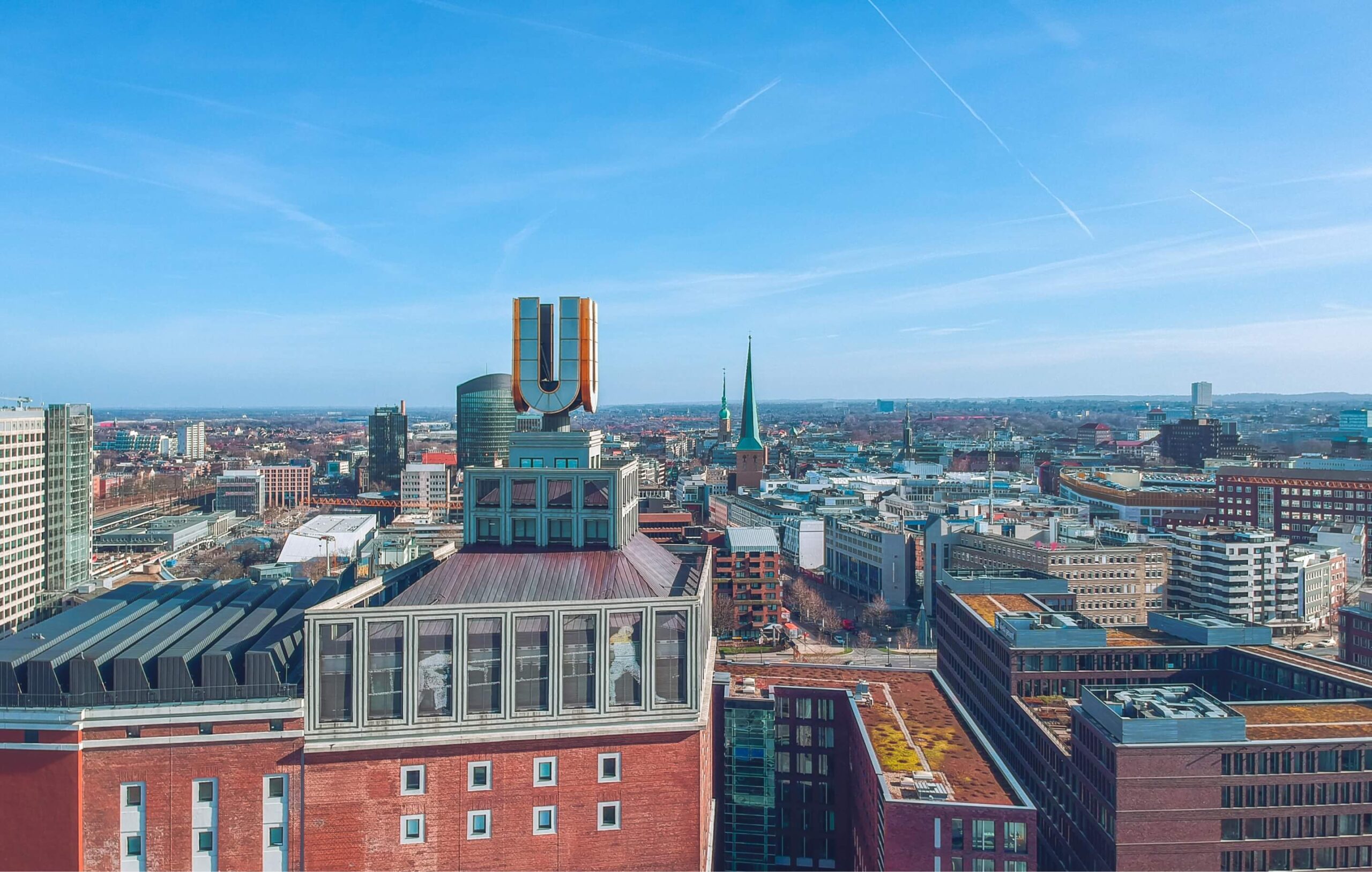 Luftaufnahme von Dortmund mit Fokus auf den Dortmunder U-Turm. Der Turm mit seinem markanten goldenen "U" dominiert das Stadtbild. Im Hintergrund erstreckt sich die Skyline von Dortmund mit verschiedenen Hochhäusern und Gebäuden. Rote Backsteinbauten sind im unteren Bildbereich zu sehen. Der Himmel ist blau mit leichten Wolken.